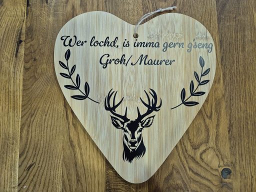 Herzförmiges Holzschild mit einem Hirschkopf und dem Schriftzug „Wer lacht, ist immer gern geseh'n“.