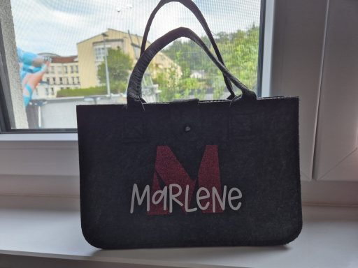 Schwarze Tasche mit dem Namen "Marlene" in bunten Buchstaben auf einem Fensterbrett.