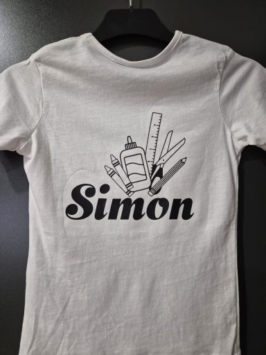 Ein weißes T-Shirt mit dem Namen "Simon" und verschiedenen Zeichenutensilien.